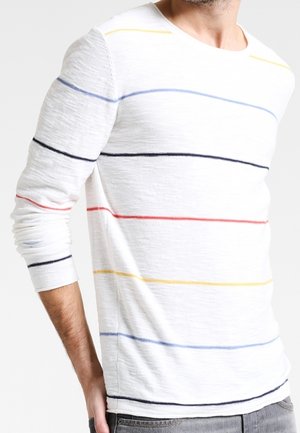 Homme portant une chemise blanche à manches longues avec de fines rayures horizontales en noir, rouge, bleu et jaune, associée à un pantalon gris.