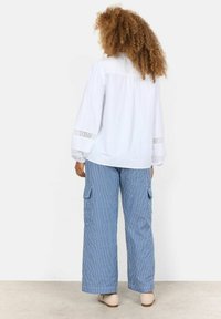 Blusa bianca con pizzo sui polsini, abbinata a pantaloni cargo ampi a righe blu e bianche con tasche laterali.