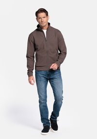 Bruine zip-up jas met hoge kraag, gecombineerd met blauwe jeans en zwarte schoenen. Casual pasvorm met ribbelmanchetten en een gladde textuur.
