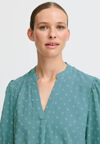 Blusa teal con scollatura a V, caratterizzata da piccole pois ricamati e leggere maniche a palloncino. Tessuto leggero con una finitura liscia.