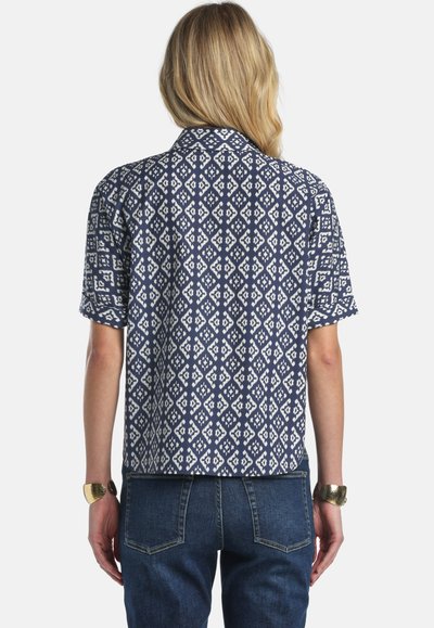 Moteris su blondiniais plaukais, dėvinti trumpomis rankovėmis tamsiai mėlyną ir baltą marškinėlį su rombo raštu bei mėlynas džinsus, matoma iš nugaros.