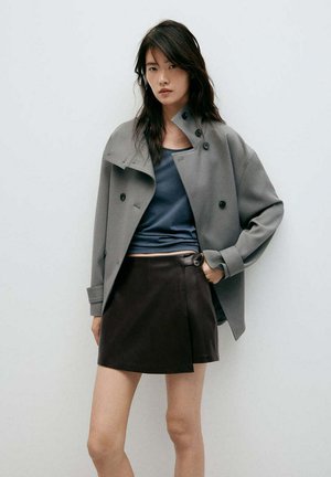 Mujer con cabello largo y oscuro con una chaqueta gris extragrande, top azul y minifalda negra, de pie frente a una pared lisa y clara.