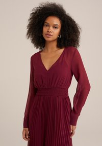 Burgundy pleated dress with sheer long sleeves, deep V-neck, and smocked waist detail, made from lightweight fabric for fluid movement.
