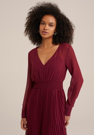 Burgundy pleated dress with sheer long sleeves, deep V-neck, and smocked waist detail, made from lightweight fabric for fluid movement.