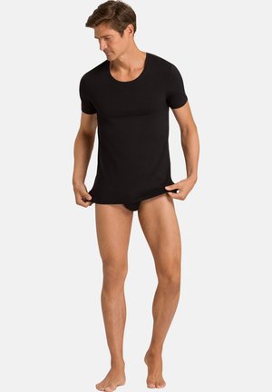 Man draagt een nauwsluitend zwart shirt met korte mouwen en zwarte slip, houdt de zoom van het shirt vast en kijkt naar links omlaag.