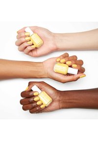 Trois mains exhibent du vernis à ongles jaune, montrant différentes carnations. Le flacon de vernis a un design carré et un capuchon blanc.