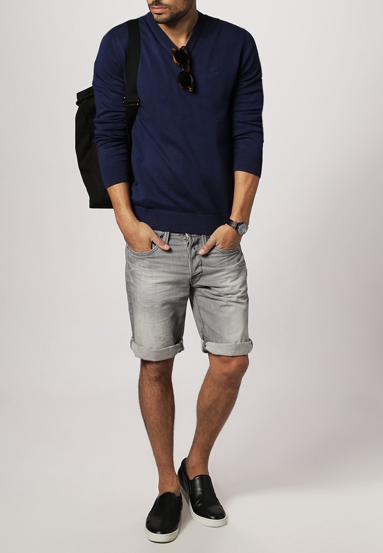 Granatowy sweter v-neck, szare, podwinięte dżinsowe szorty oraz czarne buty typu slip-on. Akcesorium: czarna torba na plecy, okulary przeciwsłoneczne.