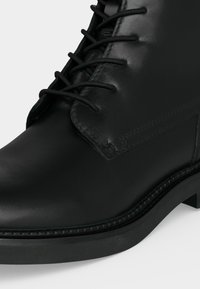 Gros plan d'une bottine noire en cuir avec lacets, coutures visibles et bout arrondi lisse sur un fond clair.
