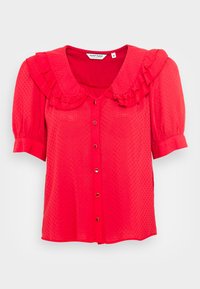 Blusa rossa con un motivo a pois testurizzato, colletto volant e maniche corte a palloncino; presenta una chiusura frontale con bottoni e dettagli dorati.