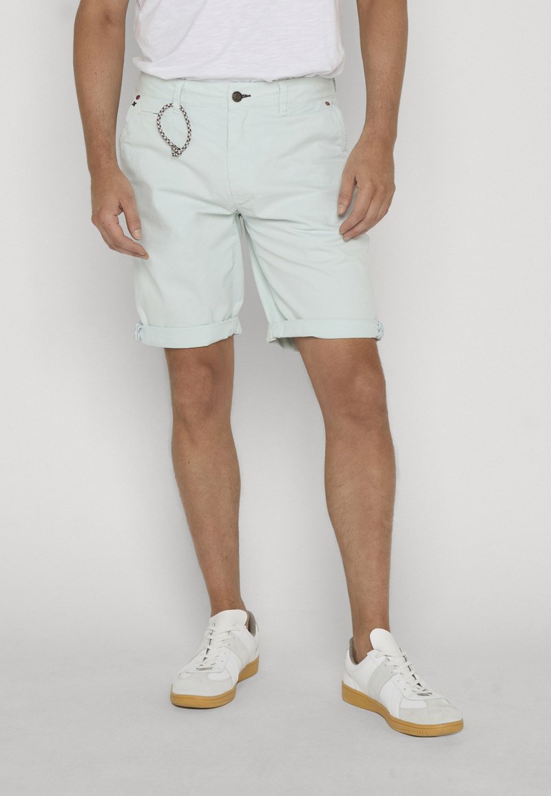Homme portant un short vert menthe clair à ourlet retroussé, des baskets blanches à semelles en gomme, et un t-shirt blanc, debout devant un fond uni.