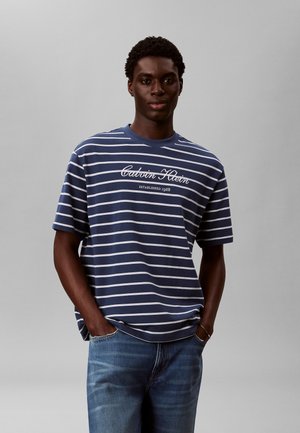 Mand iført blå og hvide stribede Calvin Klein t-shirt og blå jeans, stående med hænderne i lommerne mod en ensfarvet grå baggrund.
