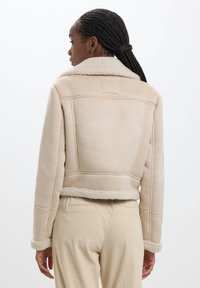 Veste en fausse fourrure beige avec un col doux, une coupe structurée, des coutures contrastées et une doublure pelucheuse aux poignets et à l'ourlet.