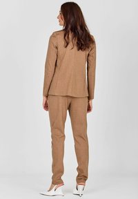 Bram jezni kostim z rjavimi pinstripi, ki vključuje krojen plašč in usklajene ožje hlače, v kombinaciji z belimi visokimi petami.