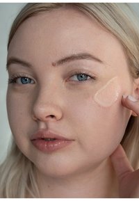 Modelo mostrando un gel translúcido aplicado en la mejilla. Presenta piel clara, cabello rubio lacio y un color de labios natural.