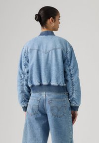 Γυναίκα με μαλλιά σε κότσο, φοράει ελαφρύ μπλε τζιν μπουφάν και ψηλόμεσα τζιν παντελόνια, γυρισμένη προς τα πίσω μπροστά σε απλό φόντο.