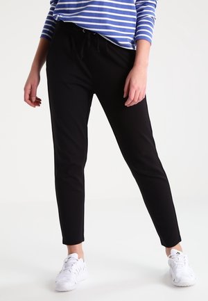 Osoba ubrana w czarne zwężane spodnie, białe trampki oraz niebiesko-biały, paskowany, długi rękaw, stojąca na jednolitym tle.