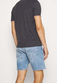 Un homme portant une chemise à manches courtes gris foncé avec des bordures roses et un short en denim bleu clair, vu de dos sur fond blanc.