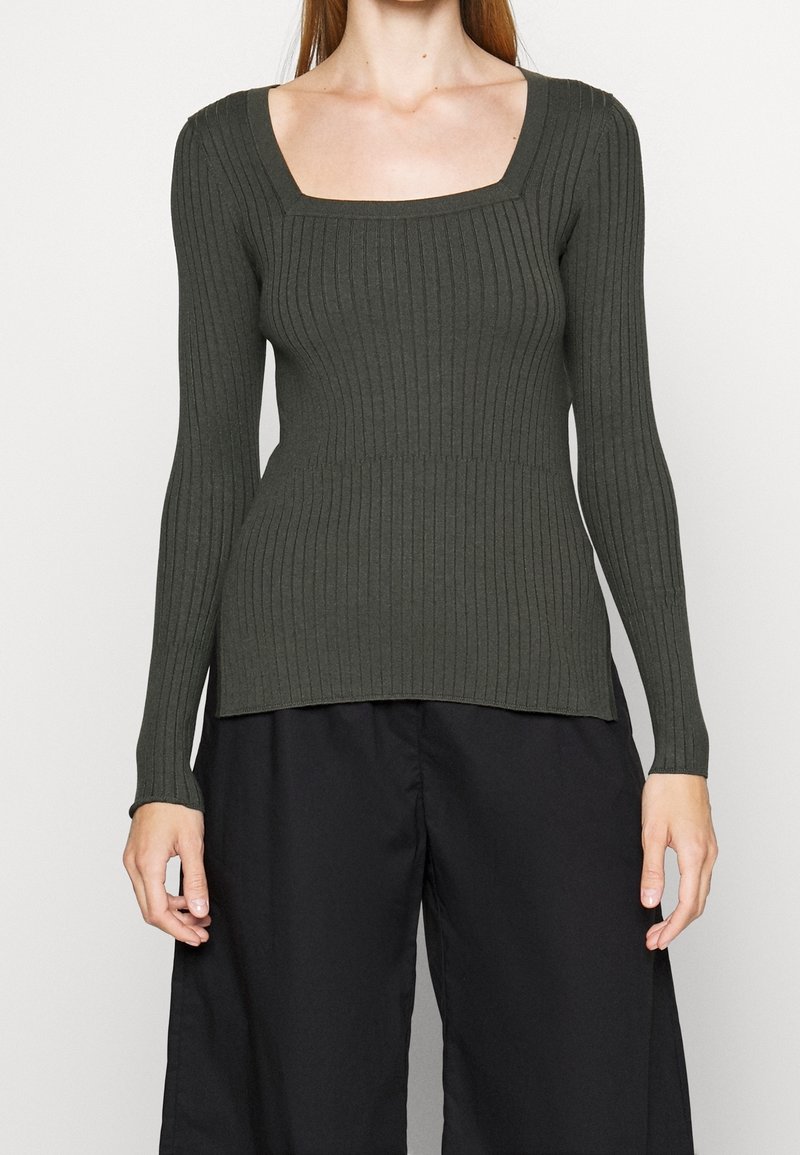 Eine Frau trägt einen figurbetonten, langärmeligen, gerippten dunkelgrünen Pullover mit einem quadratischen Ausschnitt und lockeren schwarzen Hosen, fotografiert von Hals bis Hüften.