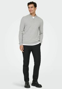 Jeune homme debout les mains dans les poches, portant un pull zippé gris clair, un pantalon noir et des chaussures noires sur fond blanc.