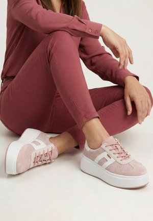 Kobieta ubrana w różowe spodnie i bluzkę z długim rękawem siedzi po turecku, prezentując różowo-białe sneakersy na platformie.