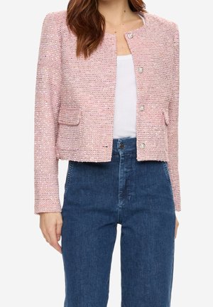 Femme portant une veste texturée rose clair boutonnée sur un haut blanc, associée à un jean bleu foncé taille haute, raccourci à la taille.