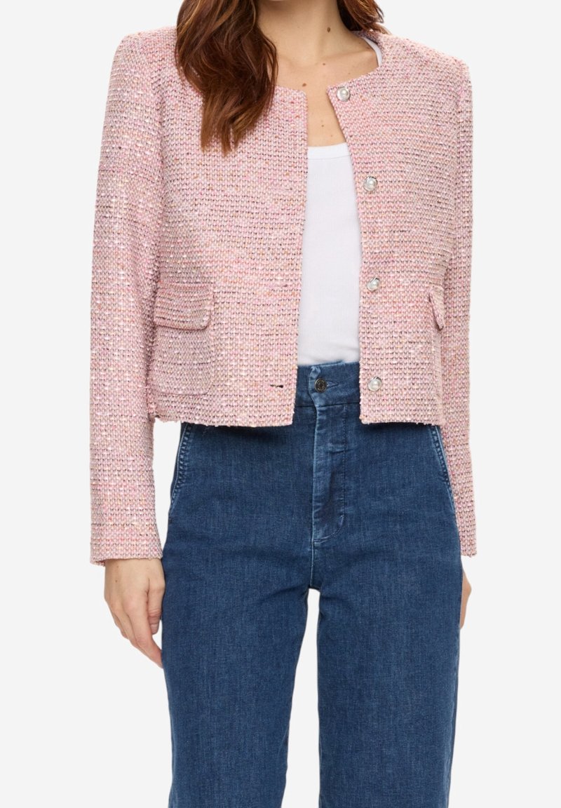 Femme portant une veste texturée rose clair boutonnée sur un haut blanc, associée à un jean bleu foncé taille haute, raccourci à la taille.