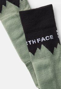 Calcetines hasta la rodilla en verde y negro, con un patrón de montañas en la mitad inferior y el logo "THE NORTH FACE" en blanco en el puño.