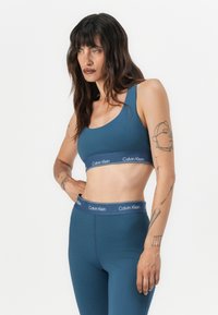 Plavi sportski grudnjak i odgovarajuće tajice s trakama s logotipom Calvin Klein. Glatki materijal, tijesno pristajuci dizajn i bez vidljivih uzoraka ili tekstura.
