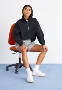 Femme assise sur une chaise de bureau orange, portant un sweatshirt noir à fermeture éclair quart, un short gris, des chaussettes blanches et des baskets blanches sur un tapis bleu.