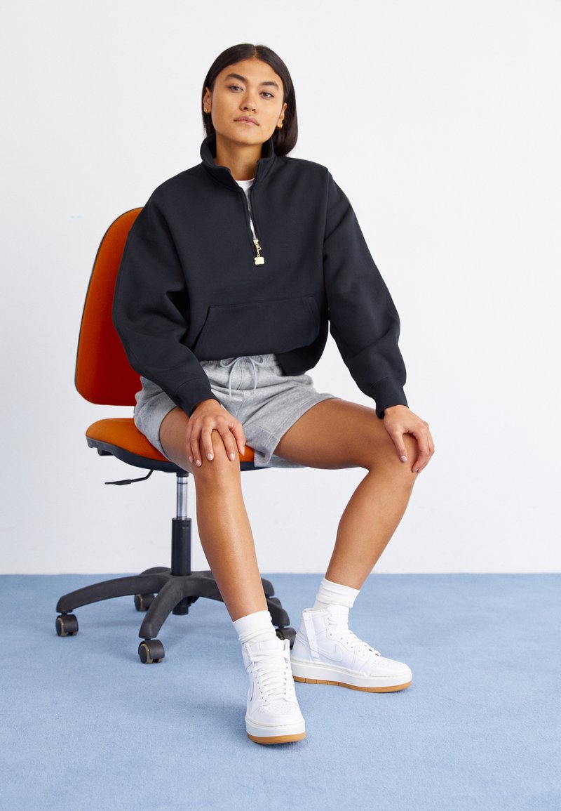 Frau sitzt auf einem orangefarbenen Bürostuhl, trägt einen schwarzen Quarter-Zip-Pullover, graue Shorts, weiße Socken und weiße Turnschuhe auf einem blauen Teppich.