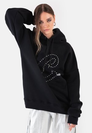 Kobieta z długimi włosami, ubrana w czarną bluzę z kapturem zdobioną ćwiekami oraz srebrne spodnie cargo, pozująca z jedną ręką za głową.