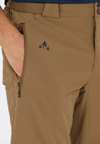 Käsi ruskeiden housujen taskussa, joissa on vetoketju ja musta kolmionmuotoinen logo lähellä etutaskua.