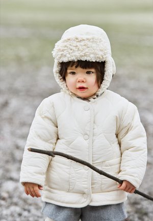 White quilted jacket with a fluffy cream hood, worn by a child holding a stick. Background is blurred, suggesting an outdoor setting.
