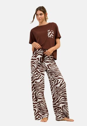 Žena stojaca naboso, oblečená v hnedej krátkosrstej košeli s vreckom so zvieracím potlačou a širokými nohavicami s rovnakým zvieracím vzorom.