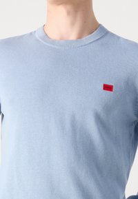 T-shirt a manica corta di colore azzurro chiaro, realizzato in tessuto morbido, con una piccola toppa con logo rosso nella zona sinistra del petto. Design semplice e aderente.