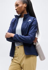 Γυναίκα με μακριά μαλλιά φοράει ένα ναυτικό σακάκι και μπεζ παντελόνι, κρατώντας ένα ανοιχτό γκρι παγούρι νερού από τη λαβή του πίσω από την πλάτη της.