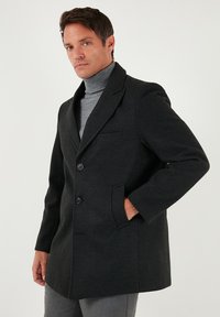 Mustast villasegust mantel, millel on sälguga krae, kaks musta nuppu, küljetaskud ja istuv lõige. Model kannab all halli kõrge kaelusega kampsunit.