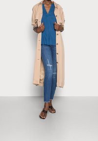 Beige Trenchcoat, blaues V-Ausschnitt-Oberteil, ausgefranste blaue Jeans und dunkelbraune Sandalen. Merkmale sind Knöpfe und ein ausgefranster Saum.