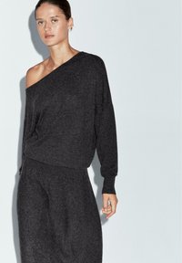 Dunkelgrauer Strickpullover mit einem schulterfreien Design, lockerer Passform und gerippten Bündchen, kombiniert mit einem passenden Rock. Weiche Textur deutlich erkennbar.