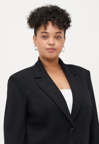 Blazer noir au design ajusté, avec des revers crantés, une fermeture à un seul bouton et un tissu texturé. Porté sur un top blanc.