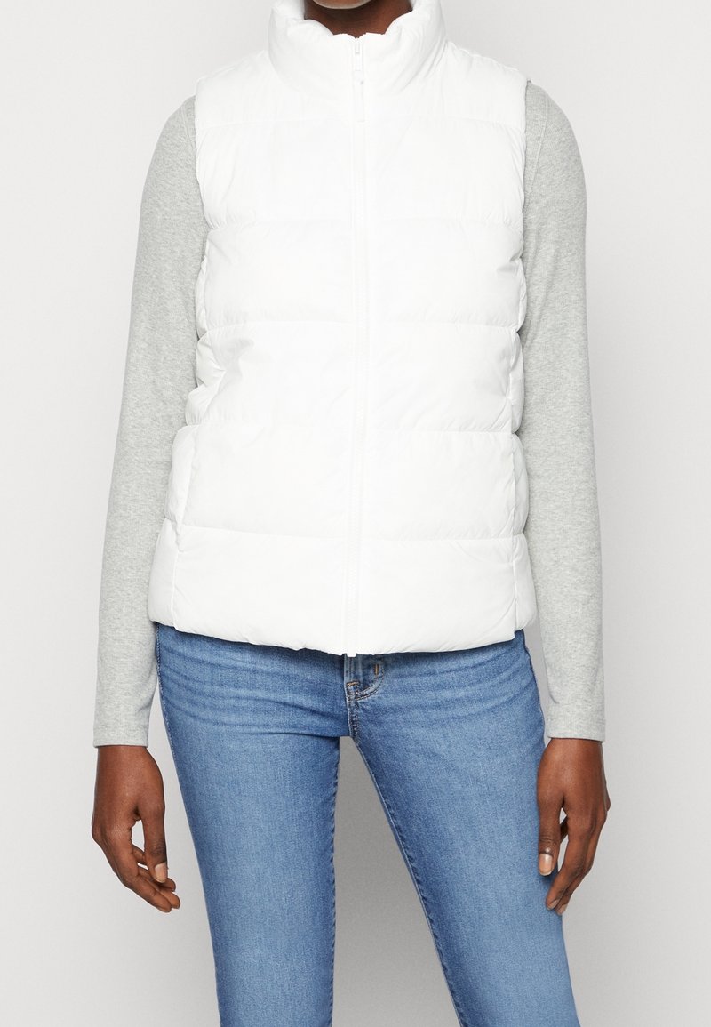 Personne portant une veste matelassée blanche sur une chemise à manches longues gris clair et un jean bleu, debout devant un fond uni.