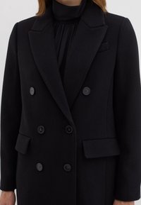 Manteau noir croisé à double boutonnage avec une finition texturée, des revers à pointes, des boutons noirs et des poches latérales. Le tissu semble chaud et robuste.