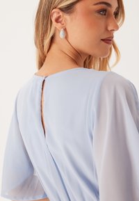 Robe bleu clair avec des manches transparentes, un col rond et un détail en forme de clé au dos. Associée à des boucles d'oreilles en forme de goutte.