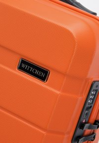 Valise rigide de couleur orange vif avec une surface texturée, comprenant une plaque de marque noire et un verrou combiné sur le côté.