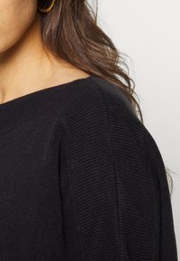 Μαύρος πλεκτός πουλόβερ με διακριτική ribbed υφή. Διαθέτει φαρδιά λαιμόκοψη και ραφές στους ώμους που πέφτουν, προσφέροντας χαλαρή εφαρμογή.