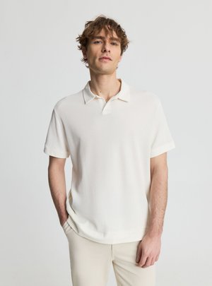 Giovane uomo con capelli ricci che indossa una polo bianca a maniche corte in maglia e pantaloni beige chiaro, in piedi con una mano in tasca.