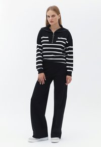 Schwarzer Pullover mit langen Ärmeln und weißen horizontalen Streifen, ausgestattet mit einem halben Reißverschlusskragen. Kombiniert mit weiten schwarzen Hosen und weißen Sneakern.