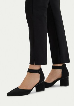 Kobieta w czarnych krótkich spodniach i czarnych zamszowych botkach na obcasie z paskiem wokół kostki oraz spiczastymi noskami na białym tle.