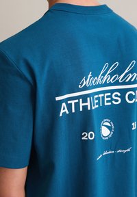 Turkusowy bawełniany t-shirt z krótkimi rękawami, z białym napisem: „stockholm” i „ATHLETES CLUB”, z dodatkowymi detalami marki poniżej.