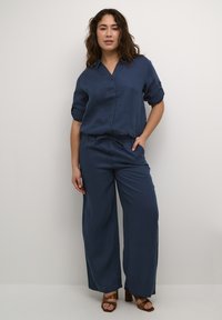 Mornarsko modra srajca iz lanu z gumbom spredaj in zavihanimi rokavi, v kombinaciji s širokimi hlačami in rjavimi bloki s peto. Gladka tekstura, sproščen kroj.